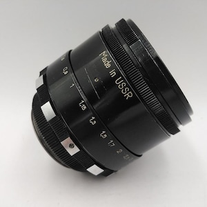 Op de afbeelding: Zwarte cameralens met zilveren markeringen en een geribbelde focusring. De lens heeft de tekst "Made in USSR" en meetindicatoren. Een vintage fotografie-accessoire.