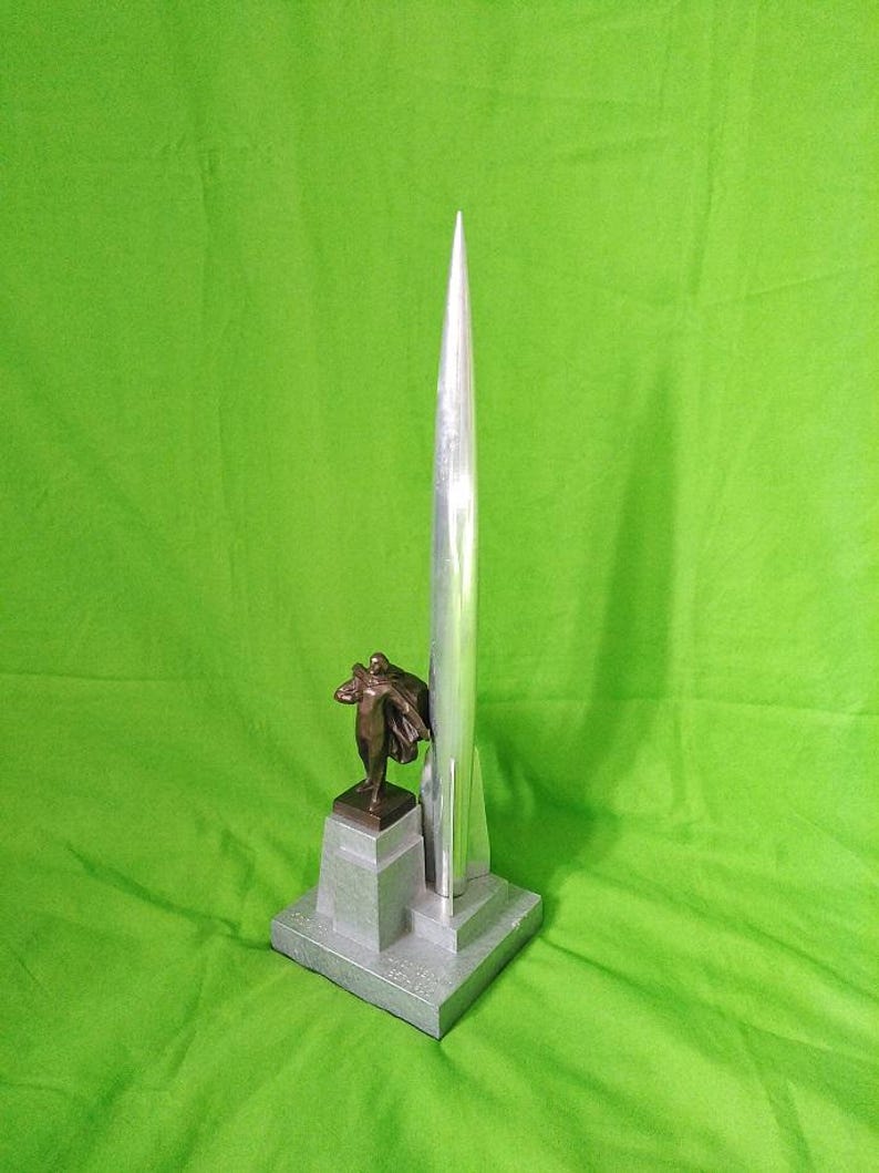 K&ouml;nnte beinhalten: Eine silberne raketenf&ouml;rmige Skulptur mit einer Bronzefigur auf einem grauen Sockel. Die Skulptur hat eine spitze Spitze und eine detaillierte Bronzefigur mit Umhang. Der Sockel ist quadratisch und hat eine strukturierte Oberfl&auml;che.