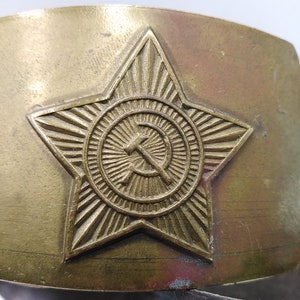 May include: A gold-toned metal belt buckle with a five-pointed star design. The star has a circle in the center with a hammer and sickle inside.