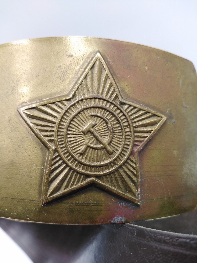 May include: A gold-toned metal belt buckle with a five-pointed star design. The star has a circle in the center with a hammer and sickle inside.