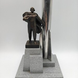K&ouml;nnte beinhalten: Eine Bronzestatue eines Mannes mit Umhang, der ein B&uuml;ndel h&auml;lt, montiert auf einem grauen, geometrischen Sockel. Hinter der Figur erhebt sich eine silberne, raketenf&ouml;rmige Struktur. Der Sockel hat eingravierten Text.