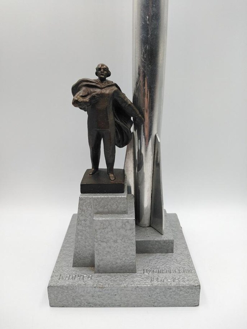 K&ouml;nnte beinhalten: Eine Bronzestatue eines Mannes mit Umhang, der ein B&uuml;ndel h&auml;lt, montiert auf einem grauen, geometrischen Sockel. Hinter der Figur erhebt sich eine silberne, raketenf&ouml;rmige Struktur. Der Sockel hat eingravierten Text.