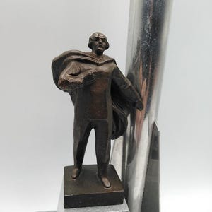 K&ouml;nnte beinhalten: Eine bronzefarbene Statue eines Mannes in einem Umhang, der auf einem rechteckigen Sockel steht. Die Statue befindet sich neben einer hohen, silberfarbenen Metallstruktur. Der Sockel steht auf einer grauen, abgestuften Plattform.