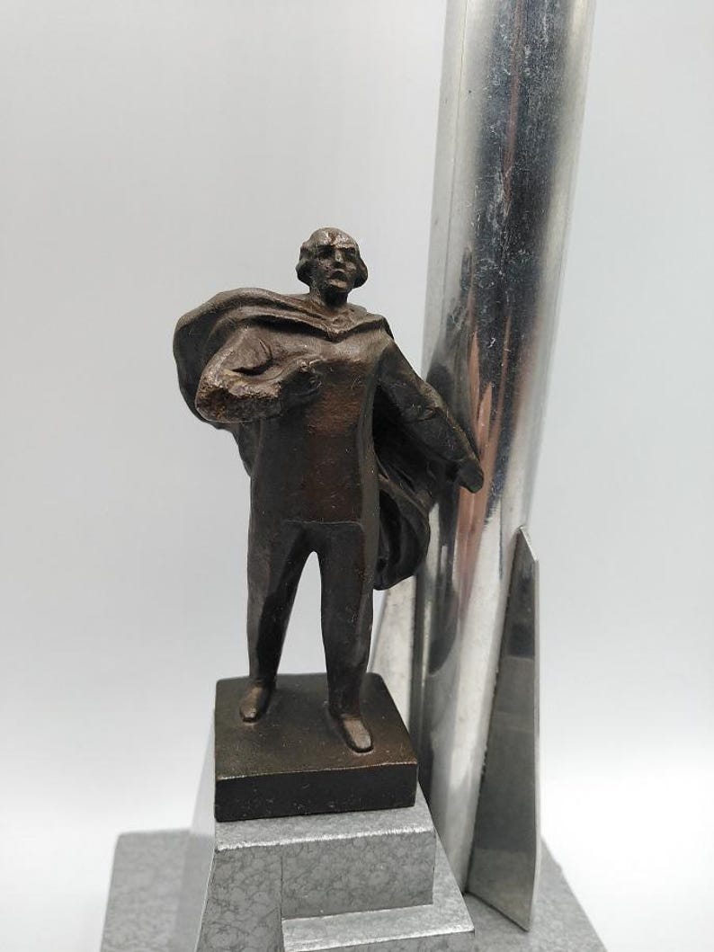 K&ouml;nnte beinhalten: Eine bronzefarbene Statue eines Mannes in einem Umhang, der auf einem rechteckigen Sockel steht. Die Statue befindet sich neben einer hohen, silberfarbenen Metallstruktur. Der Sockel steht auf einer grauen, abgestuften Plattform.