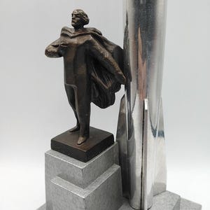 K&ouml;nnte beinhalten: Eine Bronzestatue eines Mannes mit Umhang steht neben einem silbernen, raketenf&ouml;rmigen Objekt. Die Statue steht auf einem grauen, abgestuften Sockel. Die Skulptur hat einen Art-Deco-Stil.