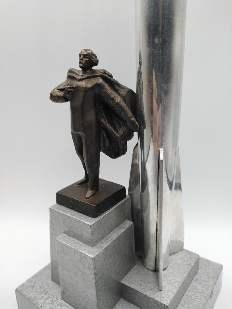 K&ouml;nnte beinhalten: Eine Bronzestatue eines Mannes mit Umhang steht neben einem silbernen, raketenf&ouml;rmigen Objekt. Die Statue steht auf einem grauen, abgestuften Sockel. Die Skulptur hat einen Art-Deco-Stil.