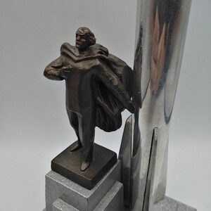 K&ouml;nnte beinhalten: Eine bronzefarbene Skulptur eines Mannes mit Umhang, der auf einem abgestuften Sockel steht. Die Skulptur wird von einem hohen, silberfarbenen Metallelement begleitet. Der Sockel hat ein graues, strukturiertes Aussehen.