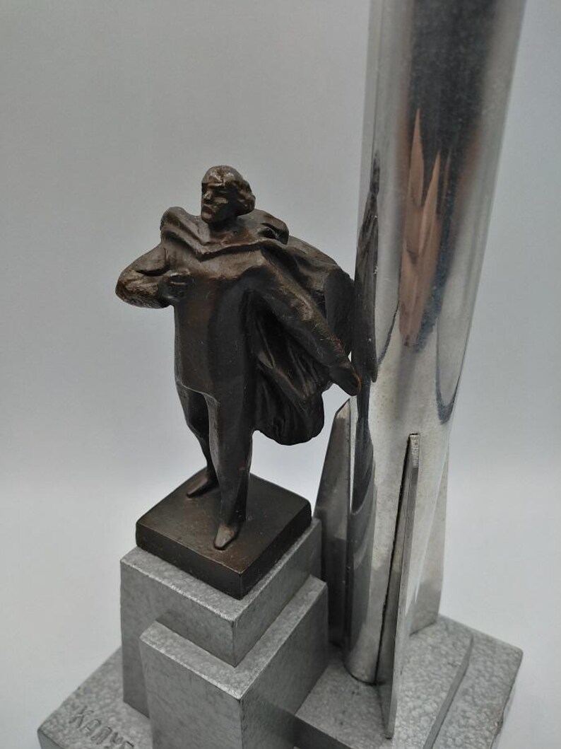 K&ouml;nnte beinhalten: Eine bronzefarbene Skulptur eines Mannes mit Umhang, der auf einem abgestuften Sockel steht. Die Skulptur wird von einem hohen, silberfarbenen Metallelement begleitet. Der Sockel hat ein graues, strukturiertes Aussehen.