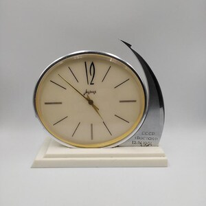 Könnte beinhalten: Eine Vintage-Schreibtischuhr mit cremefarbenem Zifferblatt, goldfarbenen Zierleisten und schwarzen Ziffern. Die Uhr hat einen silberfarbenen Akzent und einen weißen Sockel. Text auf der Uhr lautet "CCCP <BOCTOK> 12. IV. 1961".