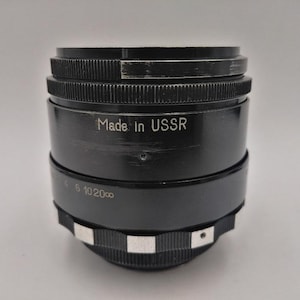 Op de afbeelding: Een zwart cameralens met de tekst "Made in USSR" tegen een witte achtergrond. De lens heeft een geribbeld ontwerp rond de randen en een zilverkleurige ring aan de basis. Het is een vintage item.