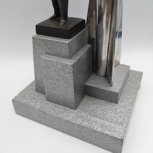 K&ouml;nnte beinhalten: Eine silberfarbene Skulptur mit geometrischem Sockel und einer Figur an der Spitze. Auf dem Sockel ist der Name "Циолковский 1857-1935" eingraviert. Die Skulptur hat eine strukturierte Oberfl&auml;che und steht vor wei&szlig;em Hintergrund.
