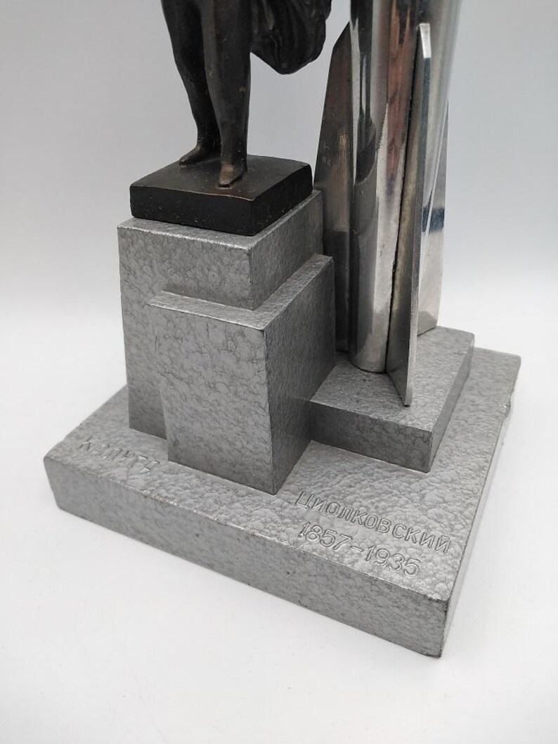 K&ouml;nnte beinhalten: Eine silberfarbene Skulptur mit geometrischem Sockel und einer Figur an der Spitze. Auf dem Sockel ist der Name "Циолковский 1857-1935" eingraviert. Die Skulptur hat eine strukturierte Oberfl&auml;che und steht vor wei&szlig;em Hintergrund.