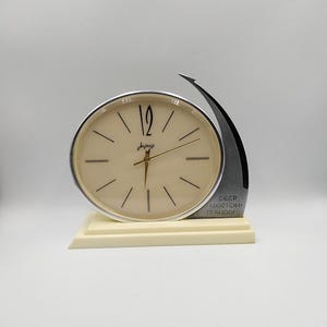 Könnte beinhalten: Eine cremefarbene Vintage-Uhr mit silberfarbenem Rahmen und schwarzen Stundenmarkierungen. Die Uhr hat goldfarbene Zeiger und steht auf einem cremefarbenen Sockel. Ein dunkelgrauer Metallakzent wölbt sich über dem Zifferblatt.