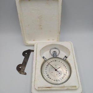May include: A vintage stopwatch with a silver-colored case and a white face, displayed in a white foam insert. The stopwatch has black numbers and hands, and is accompanied by a metal piece. The stopwatch is in an open white box.