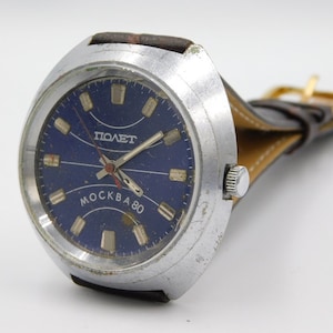 May include: A silver wristwatch with a blue face and black leather strap. The face has the words "ПОЛЕТ" and "MOCKBA 80" printed on it.