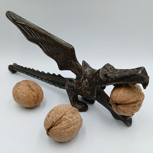 May include: A vintage metal dragon nutcracker with wings, holding a walnut in its mouth. Two additional walnuts are on the white surface.