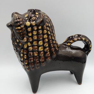 Puede incluir: Una figurita de león de cerámica marrón con una superficie texturizada y detalles amarillos. El león está de pie sobre cuatro patas y tiene la cabeza echada hacia atrás.