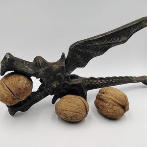 Può includere: Uno schiaccianoci a forma di drago color bronzo con un design ad ala e coda, che tiene due noci. Un'altra noce è appoggiata sotto. Lo schiaccianoci è un utensile da cucina decorativo.