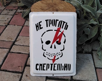 Könnte beinhalten: Weißes rechteckiges Schild mit schwarzem Text und einer Totenkopf-Grafik mit rotem Blitz. Das Schild lautet "НЕ ТРОГАТЬ СМЕРТЕЛЬНО".