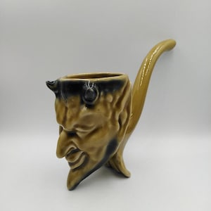 May include: A ceramic pipe shaped like a devil's head with a curved stem. The pipe is glazed in a brown and black colour.