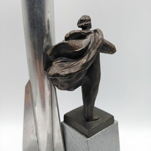 K&ouml;nnte beinhalten: Eine Bronzeskulptur einer Figur mit flie&szlig;endem Umhang, montiert auf einem quadratischen Sockel auf einem grauen Steinsockel. Links von der Figur befindet sich eine hohe, silberfarbene Metallstruktur. Die Skulptur hat einen klassischen, k&uuml;nstlerischen Stil.