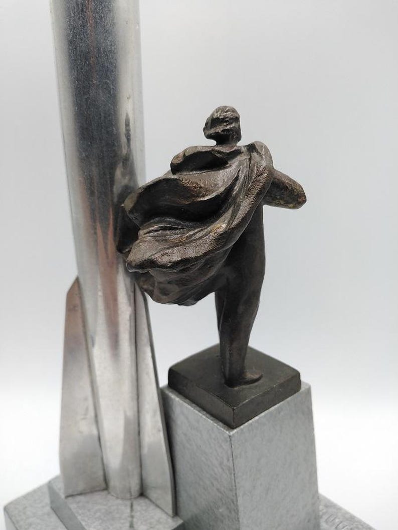 K&ouml;nnte beinhalten: Eine Bronzeskulptur einer Figur mit flie&szlig;endem Umhang, montiert auf einem quadratischen Sockel auf einem grauen Steinsockel. Links von der Figur befindet sich eine hohe, silberfarbene Metallstruktur. Die Skulptur hat einen klassischen, k&uuml;nstlerischen Stil.