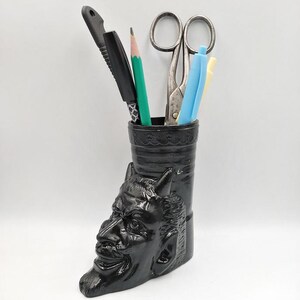 May include: Black ceramic desk organizer shaped like a devil's head with horns. It holds a pair of scissors, a pen, a pencil, and a highlighter.