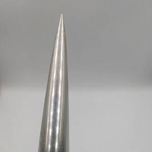 K&ouml;nnte beinhalten: Nahaufnahme eines silbernen, konischen Objekts mit einer scharfen Spitze. Die Metalloberfl&auml;che reflektiert das Licht und erzeugt Glanzlichter und Schatten. Das Objekt verj&uuml;ngt sich zu einer feinen Spitze, was auf ein Werkzeug oder einen Dekorationsgegenstand hindeutet.