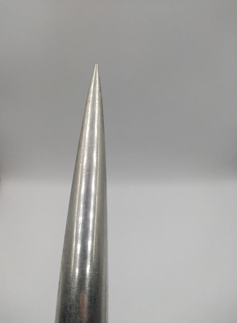 K&ouml;nnte beinhalten: Nahaufnahme eines silbernen, konischen Objekts mit einer scharfen Spitze. Die Metalloberfl&auml;che reflektiert das Licht und erzeugt Glanzlichter und Schatten. Das Objekt verj&uuml;ngt sich zu einer feinen Spitze, was auf ein Werkzeug oder einen Dekorationsgegenstand hindeutet.