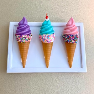 May include: Three colorful ice cream cones on a white rectangular tray. The ice cream flavors are purple, blue, and pink, each topped with sprinkles and a waffle cone. The blue ice cream has a cherry on top. The tray is against a beige background.