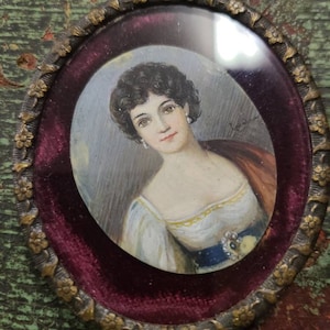 May include: An oval portrait miniature of a woman with dark hair, wearing a white dress with a blue sash and a pearl necklace. The portrait is framed in a gold-colored metal frame with a burgundy velvet backing.