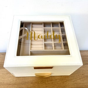May include: White jewelry box with a clear lid and gold accents. The lid has a compartment for rings and earrings. The box has a drawer and the name "Maddy" is written on the lid.
