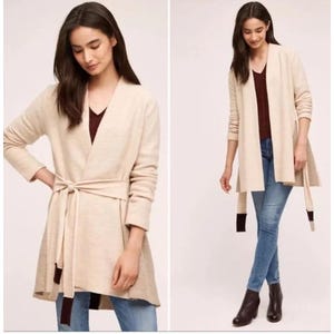 Op de afbeelding: Een beige vest met een strik in de taille en lange mouwen. Het vest heeft een V-hals en een donker bordeaux accent op de uiteinden van de stropdas. Het model draagt een blauwe spijkerbroek en zwarte laarzen.