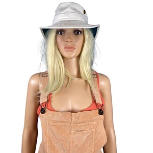 May include: A white bucket hat with a dark green underside and two bronze eyelets. The hat is worn by a mannequin wearing a peach-colored overall and a red-orange top.