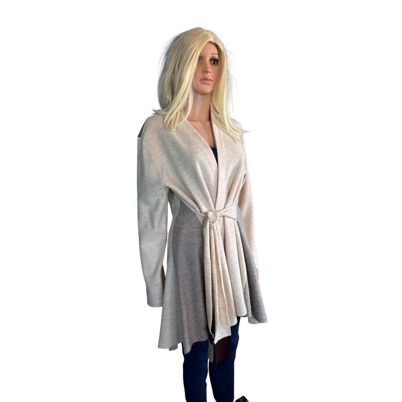 Op de afbeelding: Een beige vest met lange mouwen en een strik in de taille. Het vest heeft een gedrapeerde voorkant en een uitlopende zoom. Het model draagt een donkerblauwe broek. Het vest is gemaakt van zacht gebreid materiaal.