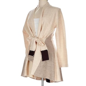 Op de afbeelding: Een beige vest met een wikkelontwerp en een striksluiting. De trui met lange mouwen heeft een colorblock rok in tinten beige en bruin. Het kledingstuk wordt op een mannequin getoond.