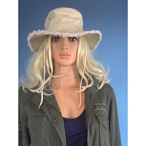 May include: A beige bucket hat with a frayed, distressed brim edge and a matching chin strap. The hat features a structured crown with small side ventilation eyelets. The fabric appears to be a sturdy cotton canvas material in a neutral light tan color.