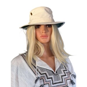 May include: A cream-colored bucket hat with a wide brim and brass eyelets. The hat has a dark green underside and is worn by a mannequin. The mannequin is wearing a white top with black and pink geometric embroidery.