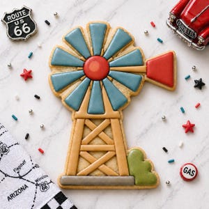 May include: A decorated sugar cookie in the shape of a windmill, featuring blue, red, and tan icing. The cookie is surrounded by Route 66 themed decorations, including a vintage car, star-shaped sprinkles, and a Route 66 sign.