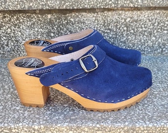 May include: A pair of blue suede clogs with wooden heels and soles. The clogs have a strap with a silver buckle and white stitching details. The heels are approximately 3 inches high.