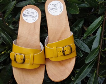 May include: A pair of yellow leather sandals with wooden soles and buckles. The sandals have a label that reads "wooden clogs natural leather SWED2WALK STOCKHOLM".