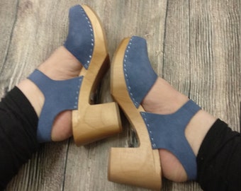 May include: A pair of blue suede clogs with wooden heels. The clogs have a peep-toe design and a strap that secures the foot. The wooden heels are a light brown color. The clogs are on a wooden surface.