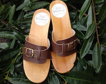 May include: A pair of brown leather sandals with a buckle closure. The sandals are made of wood and have a natural finish. The sandals have a label that says "SWED2WALK STOCKHOLM".