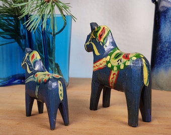 May include: Two blue wooden Dala horses with yellow, green, and red painted details. The larger horse is standing on a wooden surface. The smaller horse is standing behind the larger horse.