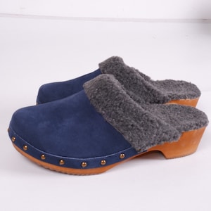 Op de afbeelding: Een paar blauwe suède klompen met een grijze imitatiebontvoering en houten zolen. De klompen hebben metalen studs rond de randen.