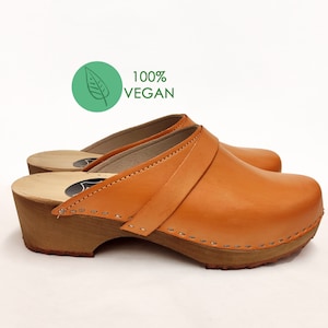 May include: A pair of orange leather clogs with a wooden heel and a stitched detail around the edge. The clogs are 100% vegan.