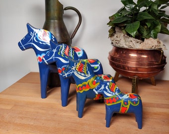 May include: Three blue Dala horses with colourful floral designs. The wooden horses are arranged in a row, decreasing in size. A copper jug and a plant in a copper pot are in the background. The horses are a traditional Swedish folk art.