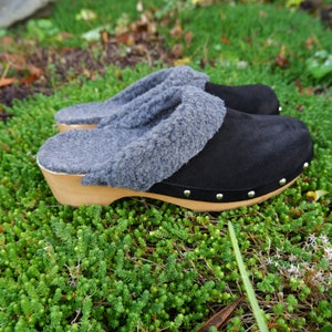 Op de afbeelding: Zwarte suède klompen met een grijze fleece voering en een houten basis. De klompen hebben metalen studs rond de rand van de basis.