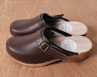 May include: A pair of brown leather clogs with a wooden sole and a buckle strap. The clogs have a white stitching detail around the edge of the leather. The brand name "Swedish Walk" is printed on a white label on the sole.