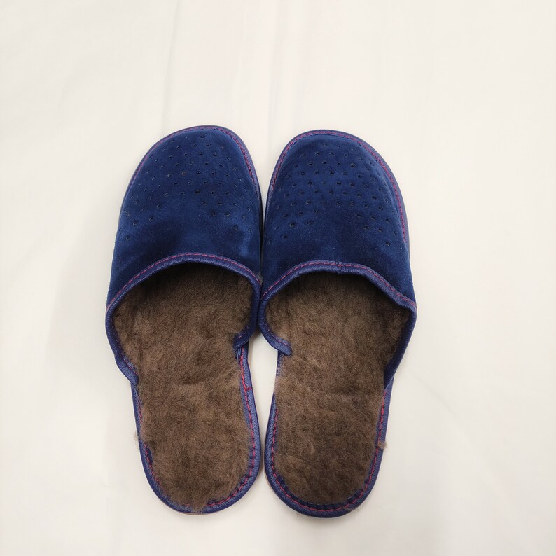 Puede incluir: Un par de zapatillas de ante azul marino con un dise&ntilde;o perforado en la parte superior. Las zapatillas tienen un interior marr&oacute;n y difuso y est&aacute;n cosidas con hilo rosa. Estas c&oacute;modas zapatillas est&aacute;n dise&ntilde;adas para uso en interiores.